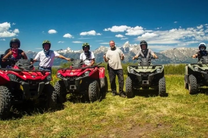 Group of five people with ATVs in a grassy area, mountains in the background.
